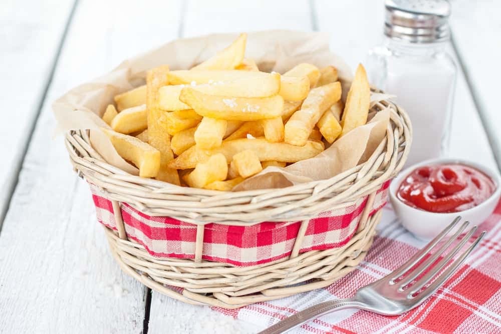 Bistro French fries in a wicker basket on white wood table with salt shaker and ketchup.