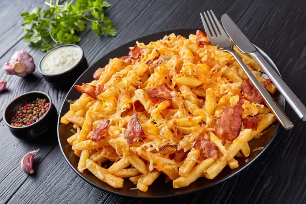 french fries with melted mix of shredded cheese and bacon served on a plate on a black wooden table with Ranch Dressing.