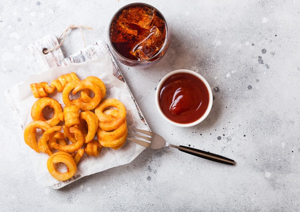 Curly fries fast food snack on wooden board with ketchup and glass of cola on kitchen background.