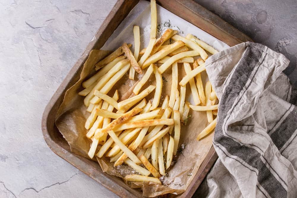 Fast food french fries potatoes with skin served with salt on baking paper in old rusty oven tray with kitchen towel over gray texture background.