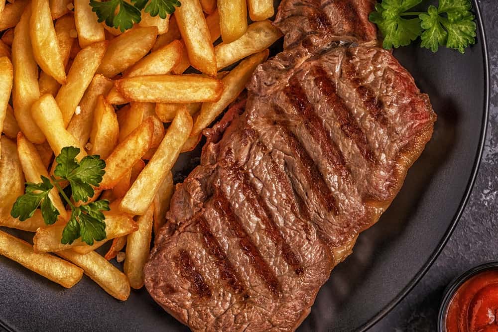 A juicy steak and its fries placed on a black plate with garnishes.