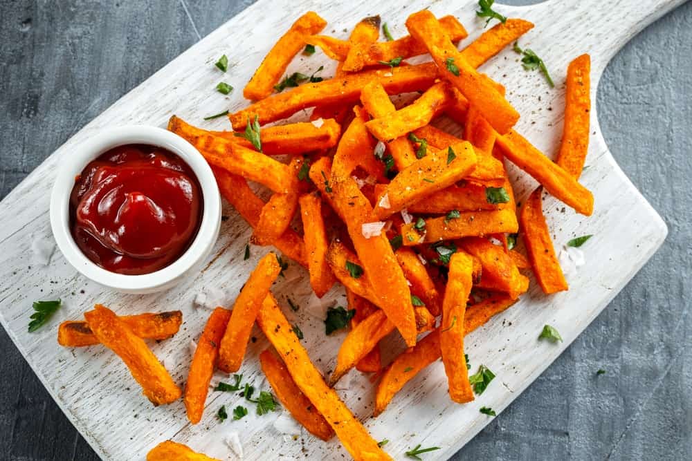 Healthy Homemade Baked Orange Sweet Potato Fries with ketchup, salt, pepper on white wooden chopping board.