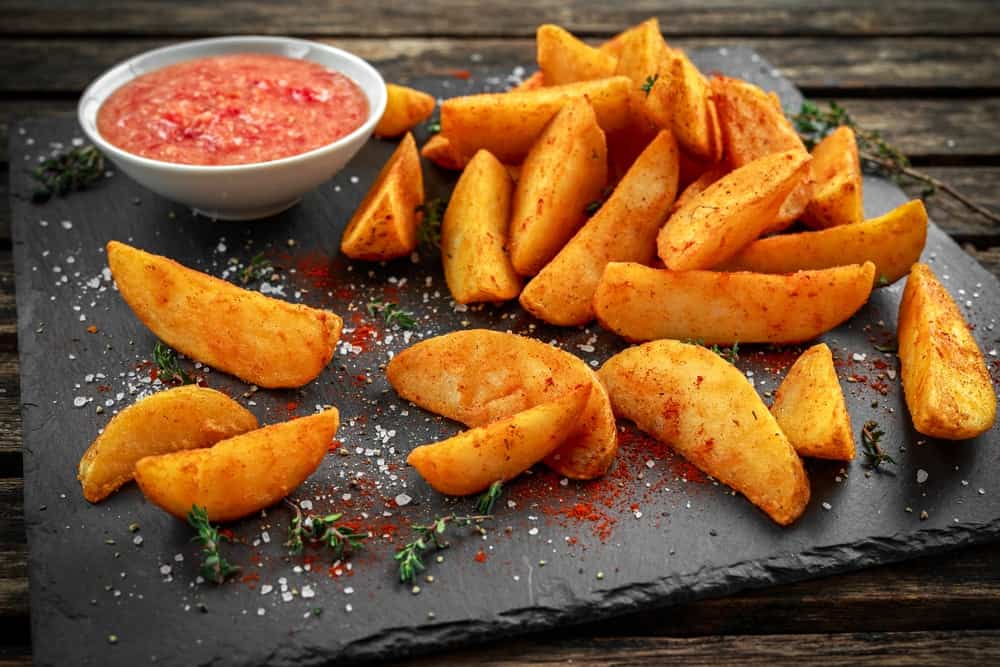 Fried potato wedges with hot salsa sauce, herbs on stone board.