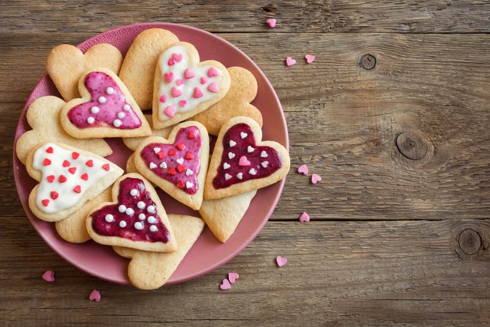 Heart cookies on a pink round plate on a wooden desk sprinkled with tiny pink hearts.