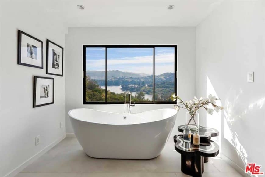 This minimalist bathroom features a freestanding bathtub with an overlooking view outside, thanks to the glass window. For a personal touch, there's an accessible black side table for plants, and some picture frames on the wall.