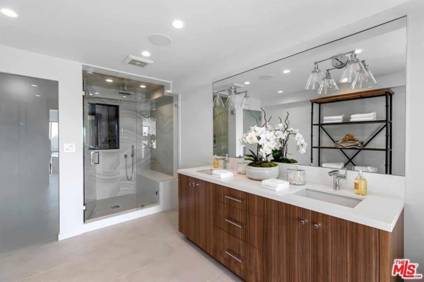 This bathroom has an Asian taste to it, particularly Japanese, with its dainty indoor flower atop a plain wooden sink and white counter, which matches the white glass-enclosed shower area with a small sitting area inside. The bathroom has wall lights that illuminate the large mirror right above the double sink.