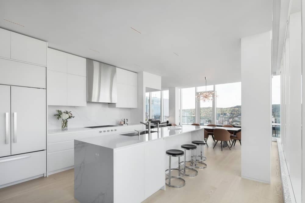 This sleek contemporary kitchen has an all-white interior that really makes it outstanding in its own right. It has a sleek white kitchen countertop paired with white drawers, and even white walls to complete the look. The island is paired with black bar stools, and just beside it is the white long dining table with brown mid-century chairs, all under a beautiful peach chandelier.