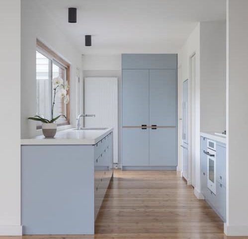 This modern kitchen has the perfect touch of simplicity to it with its wooden flooring, baby blue furniture, and white walls. The glass window by the sink also gives a great view of the outside.
