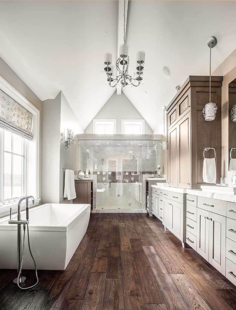 Large modern primary bathroom featuring large freestanding rectangular bathtub matches the rustic hardwood flooring. It also has wooden cabinetry and the room brightens by a chandelier.