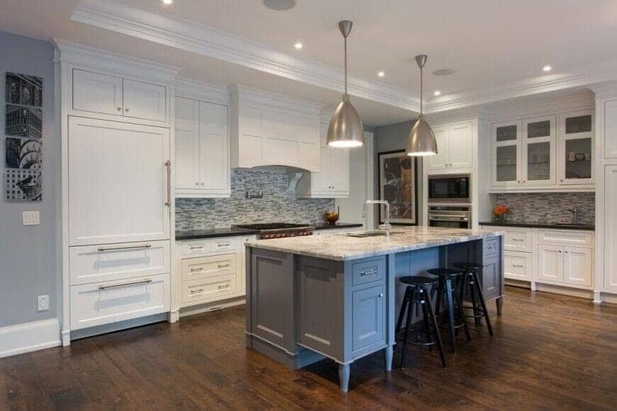 This contemporary kitchen has a beautiful classic combination of grey, white, and brown. The metallic pendant lights give a good glow over the marble and grey island with a wooden flooring underneath. The island is also surrounded by white drawers and shelves with a gold handles for a nice touch.