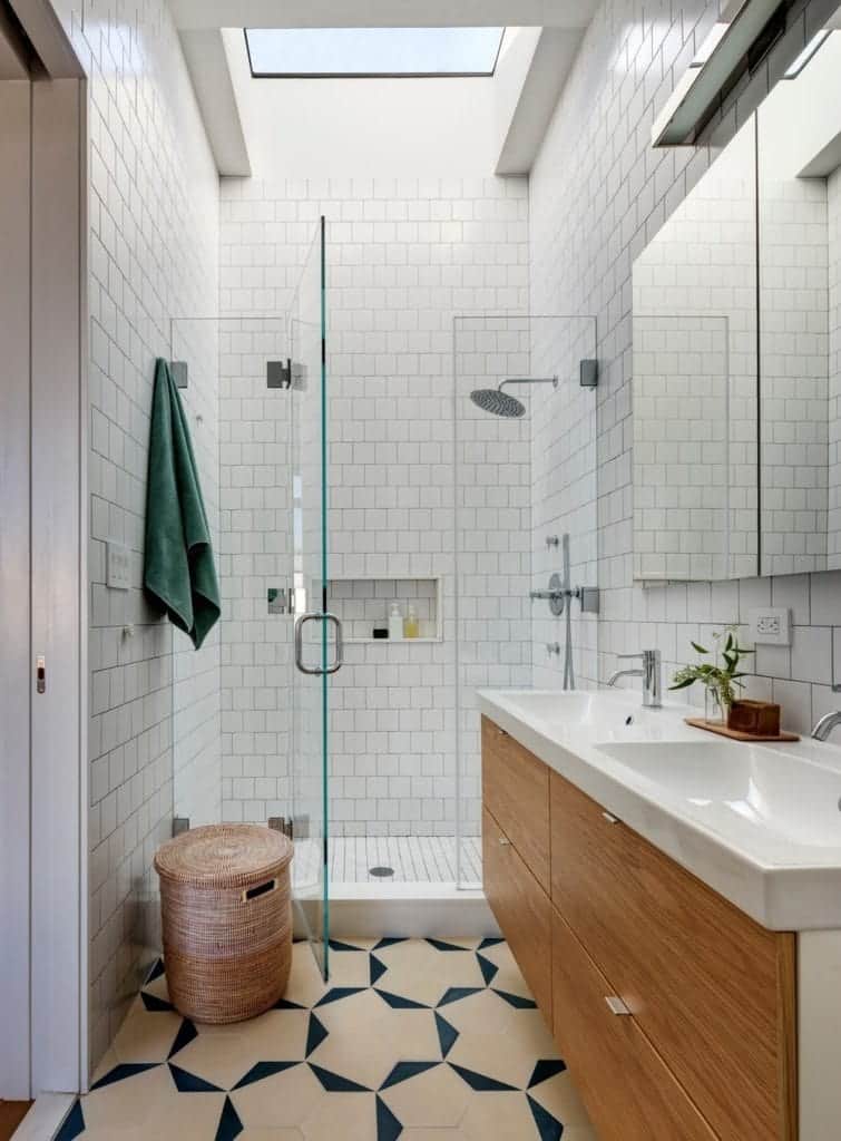 The small bathroom features wooden cabinetry and stylish carbage can. It is accompanied by a walk-in shower and a dual sink vanity against the light white marble walls.