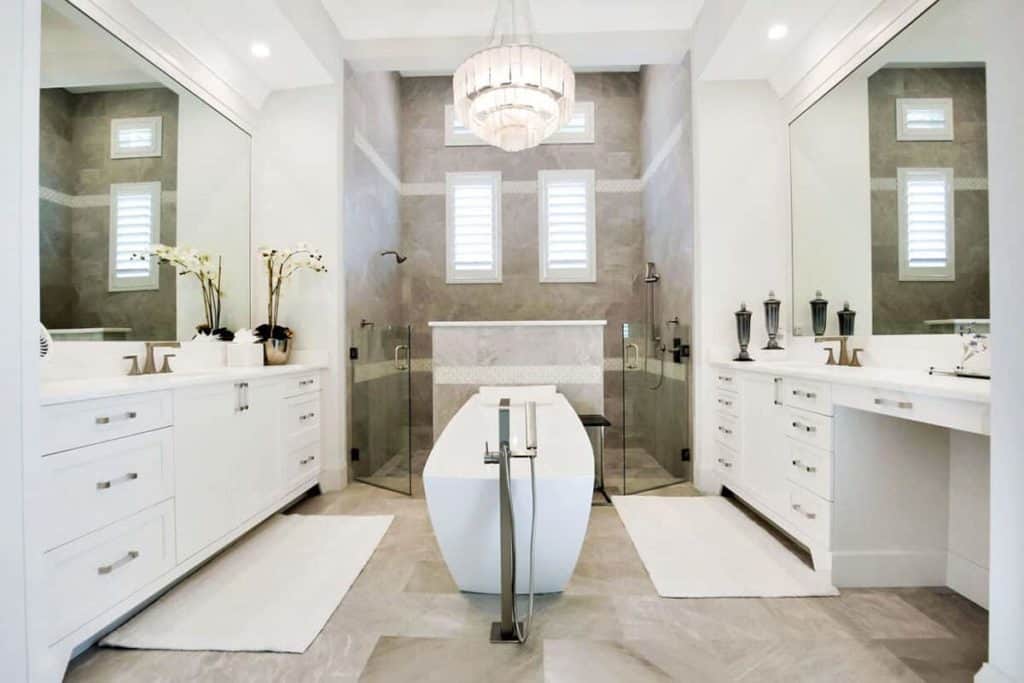 This large bathroom has a generous space for two, given its long counter with a sink on each side of the room, both with their individual bathroom essentials. Right in the middle is a white freestanding tub, with the large double shower behind it. To keep the room clean at all times, there are large white rugs on the marble tile flooring. The fancy 3-tier chandelier finishes the fancy look.
