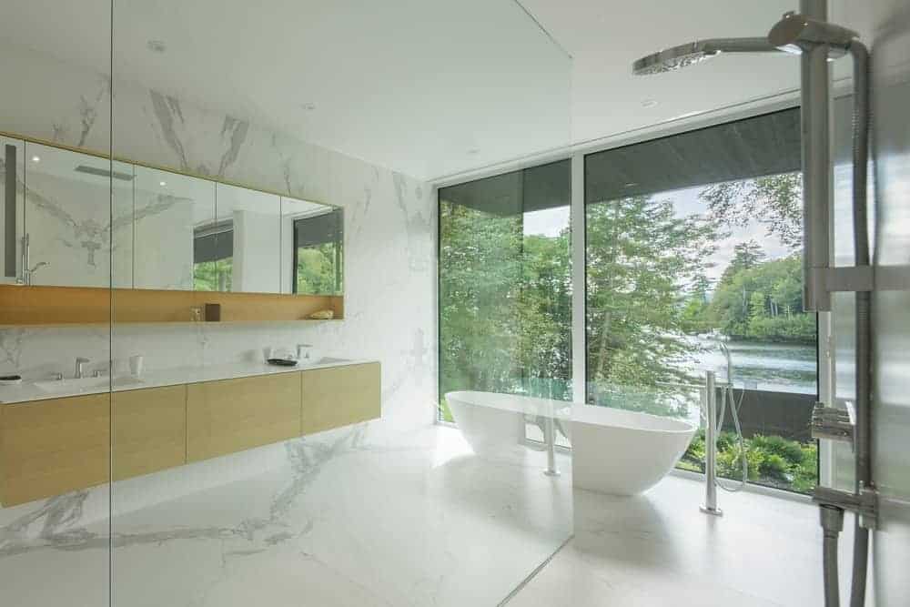 Small bathroom featuring double sink countertops match by white marble flooring. It also bathtub in the middle and a glass wall with a great view outside.