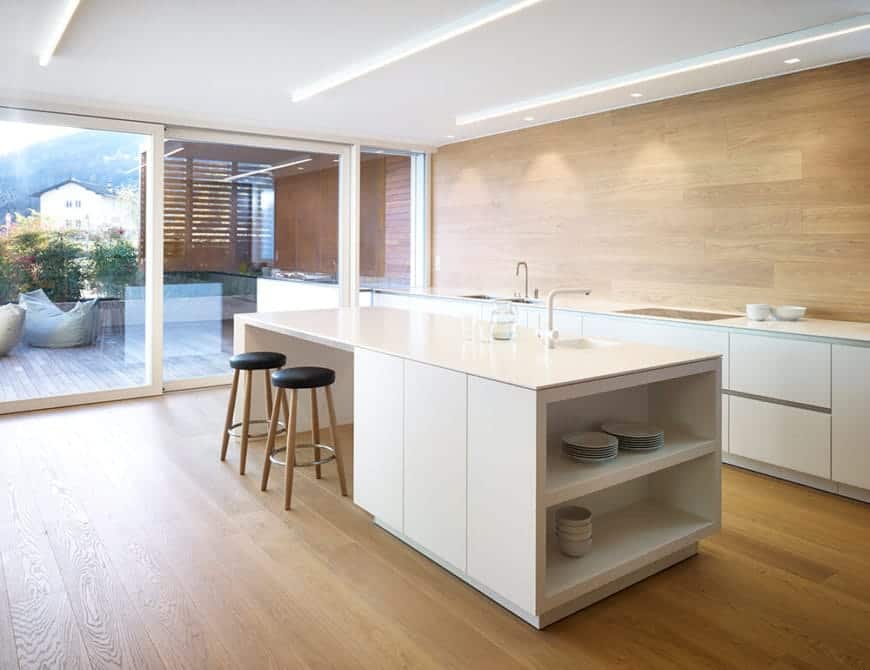 This kitchen has a minimalist design with its white island that matches the white counter. Both bars have a sink for convenient cleaning. The side of the island provides spacious shelves for plates and bowls. The wooden flooring goes well with the black and wood bar stools in place. With the floor-to-ceiling glass window, it adds a sophisticated touch to the kitchen.