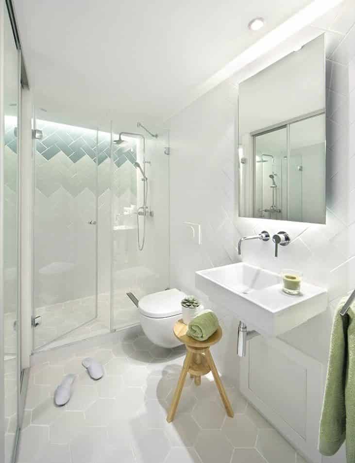 This bathroom has a nice minty feel to it with its all-white interior, balanced off with green accents. It has nice light grey hexagonal tiles that match the white floating toilet as well as the floating sink with a frameless mirror on top. The shower area has a nice medium size with lovely gradient herringbone tiles on the wall.
