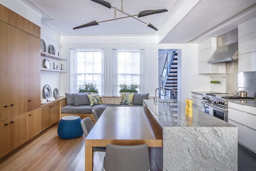 The white kitchen shares the room with a wood and grey dining area and living area for a neutral modern design. Focusing on the kitchen, it has a white counter with steel cookstove, rangehood, and oven. With the grey marble island right across, it's convenient to lay plates of food, with the 4-seater dining nook attached directly to it.