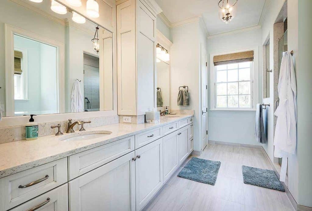 This large bathroom features a lovely double-sink counter for individual use. The blue floor rugs and towel give a pop of color in this all-white interior, including the lightings, counter, and shelves. The large mirrors are perfect for this spacious bathroom, while the window at the end of the room allows for a quick view outside.