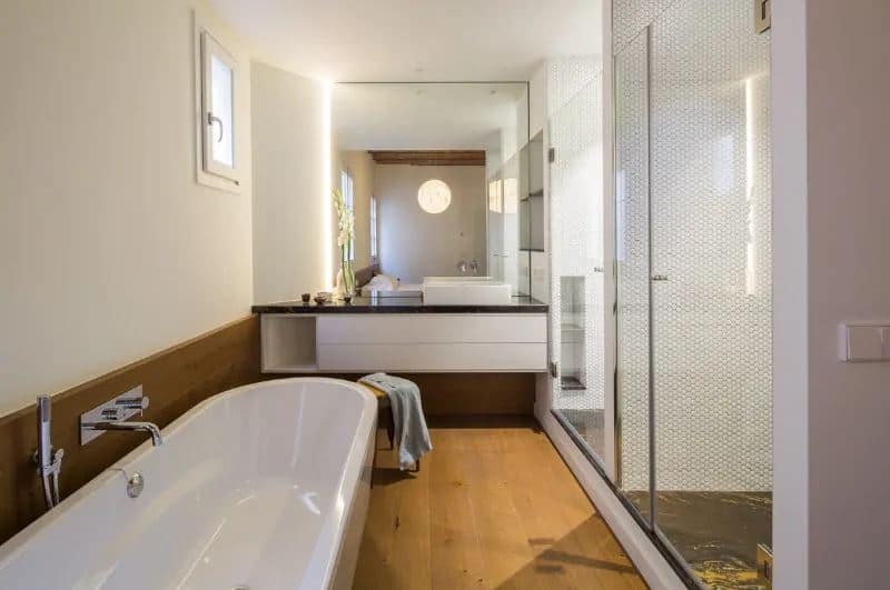 Small bathroom with freestanding bathtub on the hardwood flooring and dark countertop with a basin sink. This bathroom boasts a walk-in shower and it has mirrors that mounted on the white walls.