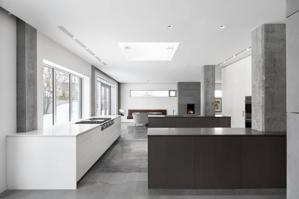 This chic minimalist kitchen features a sleek counter with a fuss-free stovetop, dark brown dining bars with grey pillars, and smooth grey marble flooring as well. It has recessed lights on the ceiling for better light distribution, and glass windows and doors for a nice outdoor view.
