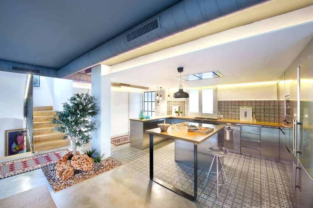 This simple Spanish kitchen features a white wall with floral patterns, which are emphasized by the yellow wall lights. It has an L-shaped counter, plus a square breakfast bar with a steel stool. All the appliances are also steel for a consistent look. This kitchen has an indoor plant at the corner, making this open kitchen look more welcoming.