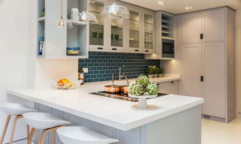 This Spanish style kitchen has a nice blue-green splashback that complements the white L-shape countertop, mirrored shelves, and translucent pendant lights.