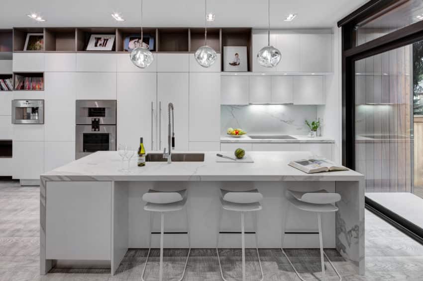 Cook up a feast for your loved ones on the wide white island covered in a marble design, which goes well with the grey flooring and the white pantries beside the sink. What makes this kitchen look more welcoming are the stack of books and picture frames lined up on the shelves. For the lighting, this kitchen has unique round pendant lights right above the counter, recessed lights, and sink lighting.