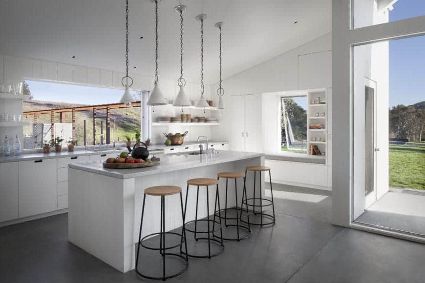 This Scandinavian style kitchen offers a refreshing vibe for your guests with its all-white interior including the island, triangular pendant lighting, door frame, walls, and drawers. There are also plenty of shelves for plates and utensils, a set of rustic-industrial bar stools, and glass windows that welcome a relaxing view from outdoors.