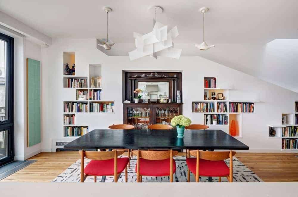 Modern dining area and wooden dark table along with wooden chairs on the rug. This room highlighting a white wall and bookshelf.