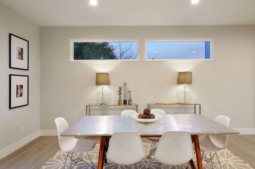 A dining room with a modern table and chairs on the rug. This room features a grey wall with great wall decors lightens up by a pair of a table lamps.