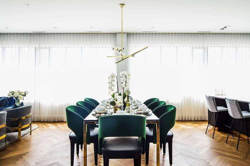 The dining room boasts an elegant theme featuring multiple glass windows blending with the white walls and hardwood flooring along with green seats.