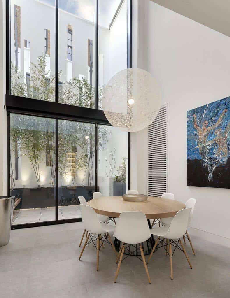 A stylish home with a wooden round table set surrounded by white walls with art design and a huge window with a pleasant view of the garden.