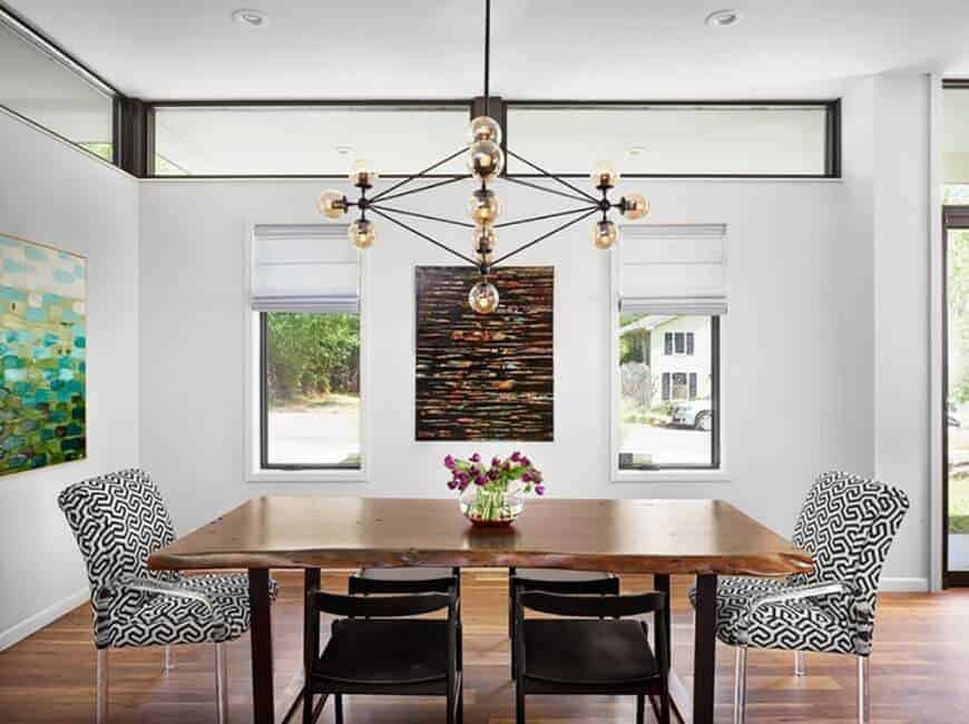 Small white dining area highlighting hardwood flooring and wooden tables with a pair of modern chairs light up by chandelier. There's a window with a view outside the house.