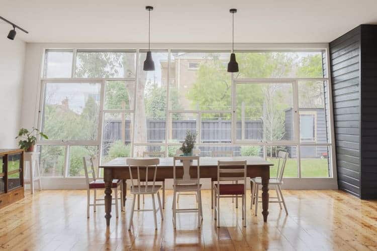 Small dining room featuring hardwood flooring and wooden tables with wooden chairs light up by hanging lights. This room also features a large window with a pleasant view outside.