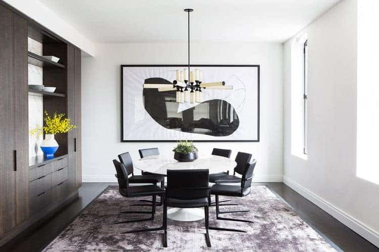 Modern dining area with round table pair with black chairs brightens by an impressive chandelier. This room has white walls and huge art.