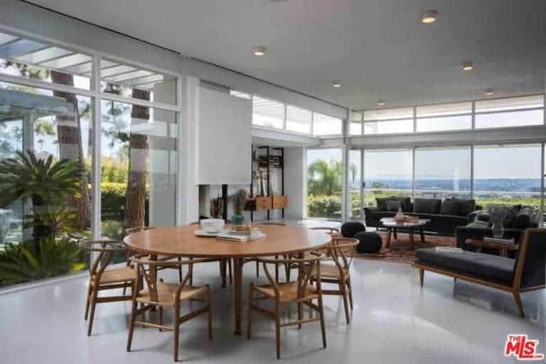 A modern house with a living space and dining area. The round wooden tables paired with wooden chairs, this room also features a glass wall and a great view outside.
