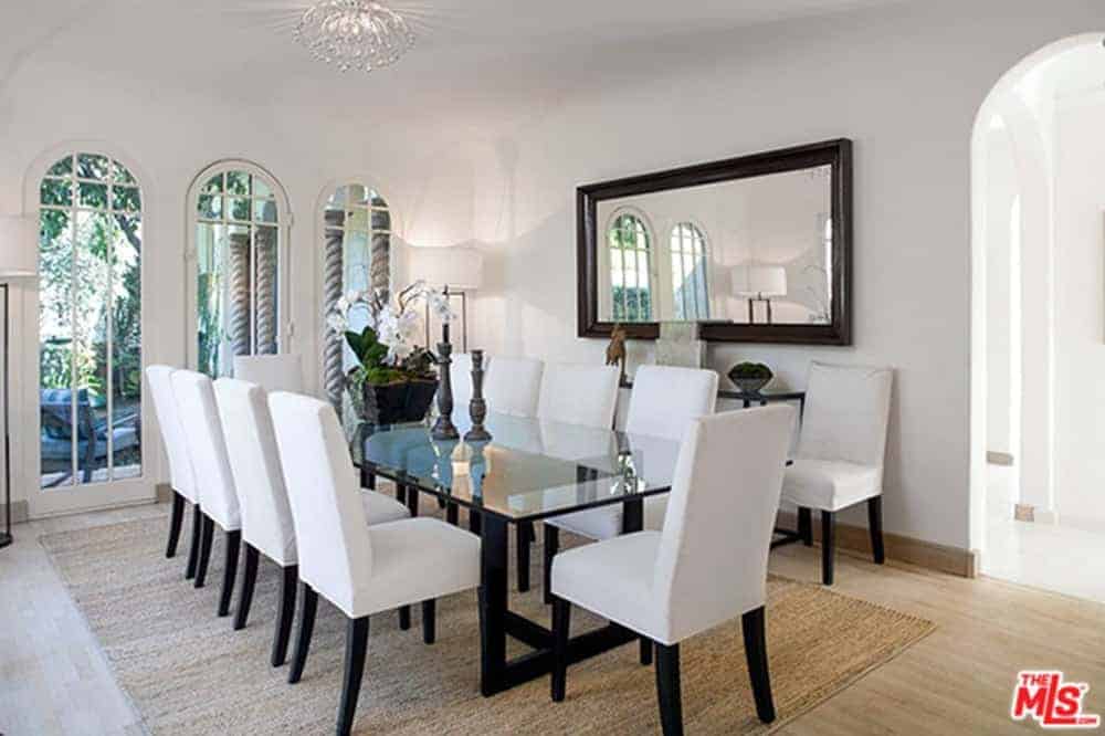 This white dining room featuring a large rectangular glass table paired with white chairs on the carpet. It also features a large window with a gorgeous garden outside.
