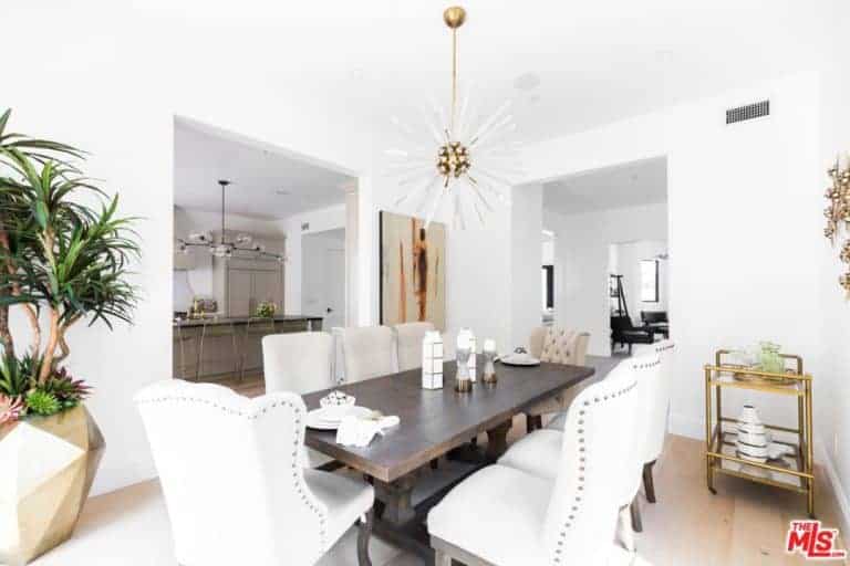 A large dining area features a wooden table mixed with modern white chairs. It also features a white wall and a white ceiling lighted by the modern chandelier.