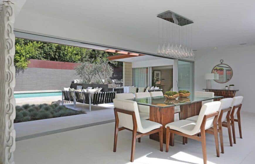 Great room with living space and dining area with glass rectangular tables with wooden chairs lighted by an elegant chandelier.