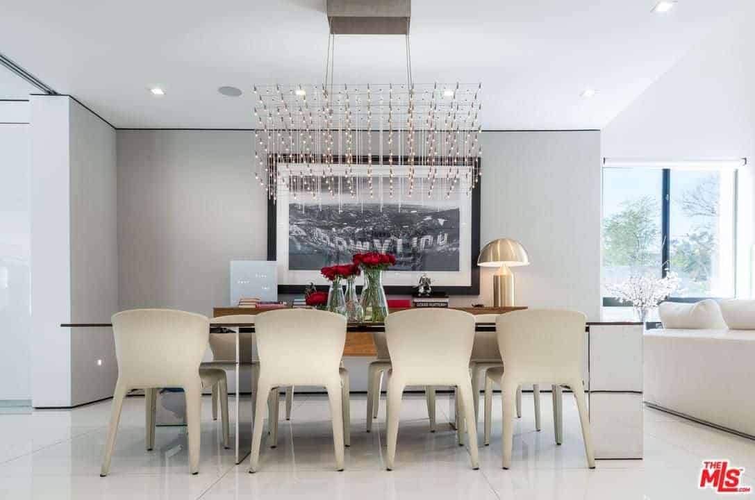 A classy dining area featuring an elegant dining table with white chairs lighted by the stunning chandelier. It also features white walls with huge wall decor.