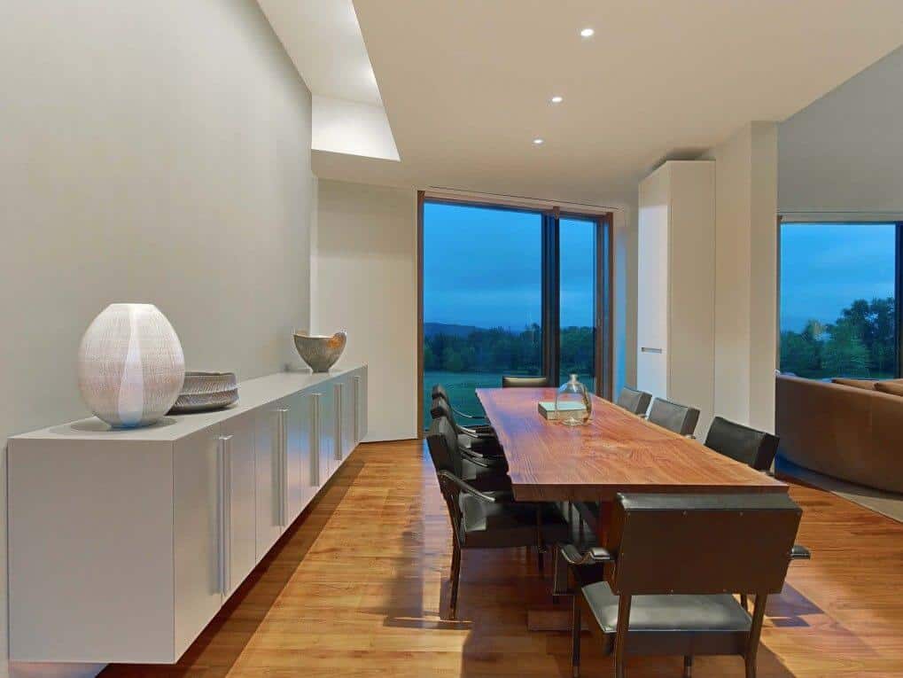 A large dining room highlighting a hardwood flooring and wooden dining table set and elegant dark seats lighted by recessed ceiling lights.