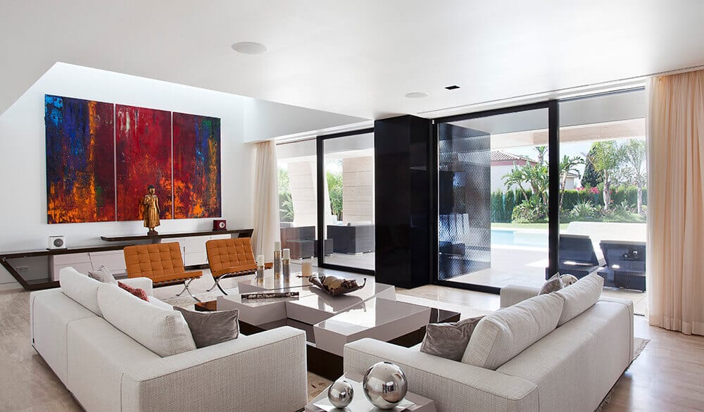A modern house with a classy living room featuring a white wall with large wall decor. This room also features a classy sofa set and a large window with a great view outside.