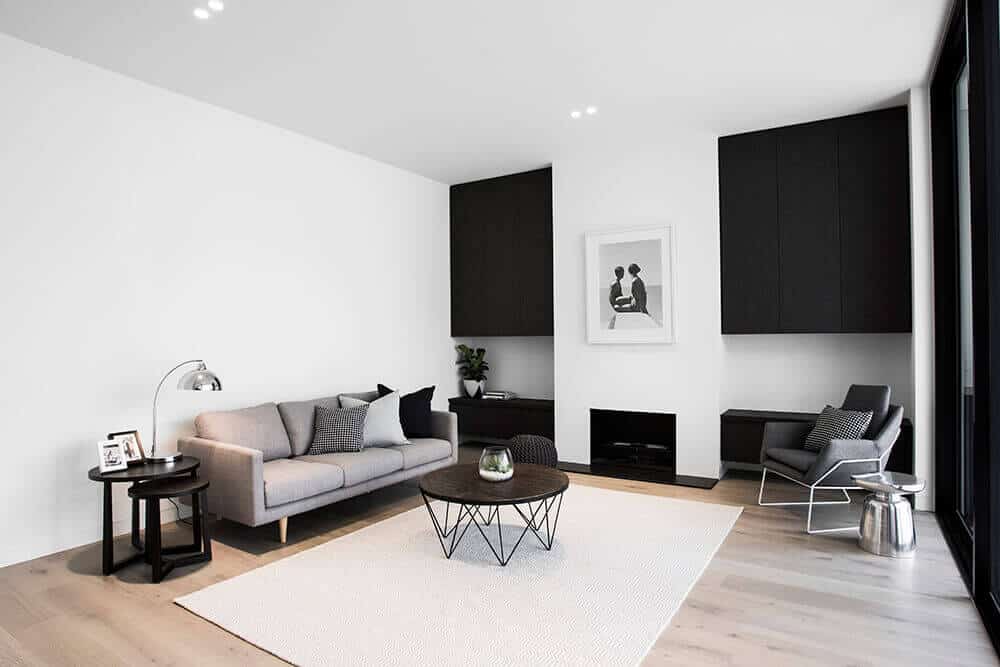Modern white living room combined with black cabinet. This room features a wooden round table on the rug and a modern couch with a table lamp beside.
