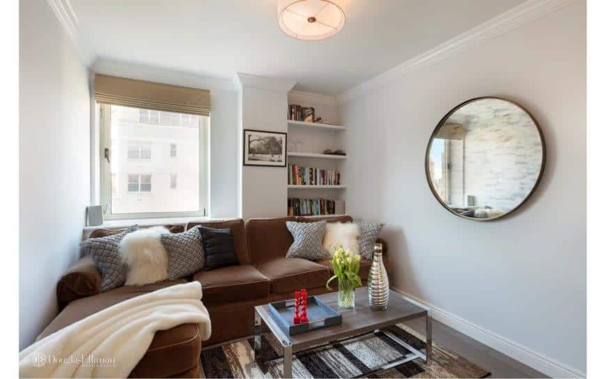 Small living room featuring an L-shaped brown sofa set. This room also has white walls with a circular mirror and bookshelf.