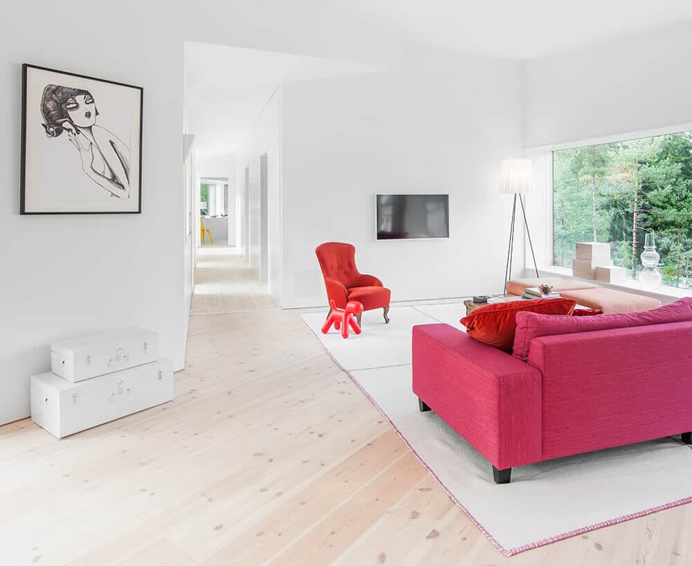 White living room with an attractive red sofa set on the carpet and also has a TV on the wall. This room has a glass wall with a great view.