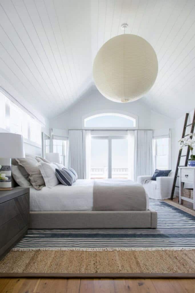 Master room highlighting hardwood flooring covered by an enormous carpet encompassed by white walls, high ceiling with immense pendant lighting and a beautiful balcony outside.
