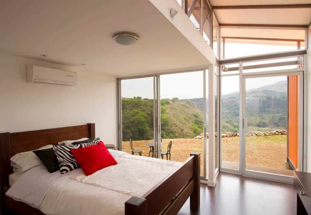 This modern master bedroom has white walls alongside a flush mount lighting. It has glass walls showing a stunning scene outside.