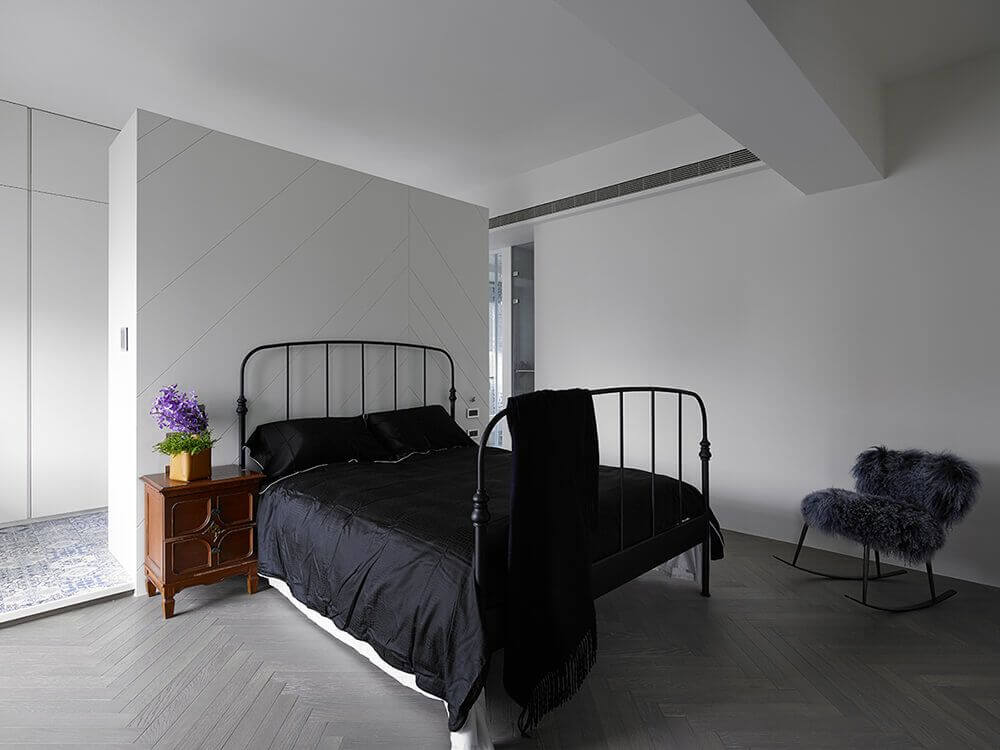 This master bedroom has herringbone-style hardwood flooring. It 's surrounded by white walls and ceiling with a cute little side table and a furry rocking chair.