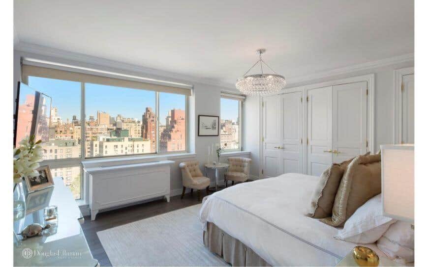 This master bedroom highlights hardwood flooring covered by an area carpet. It has a large windows with a sitting area nearby and a beautiful view outside. The room is lit by an extravagant light fixture.