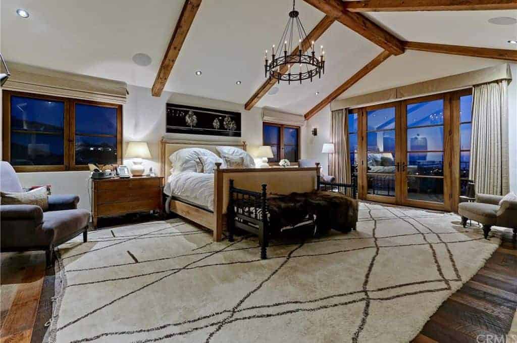 This white master bedroom with a glass door leading to a small balcony together with an elegant chandelier ceiling lights.