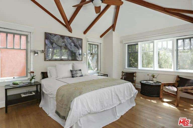This master bedroom has a comfortable bed set on the hardwood flooring, lit by two hanging lights with a couple of side tables and a great view outside.